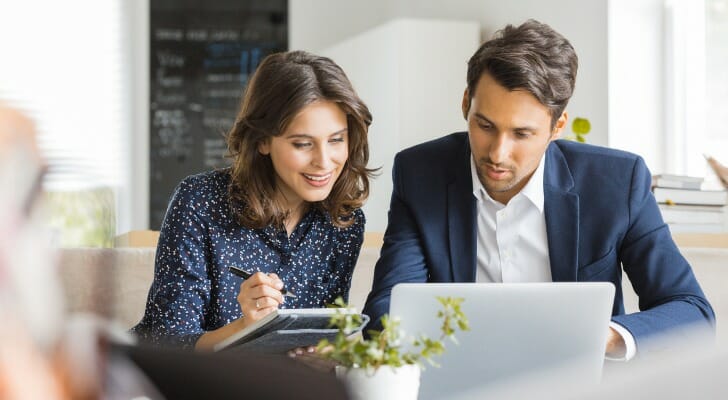 Office boy looking at a laptop while a girl behind him holds a notebook in her hand