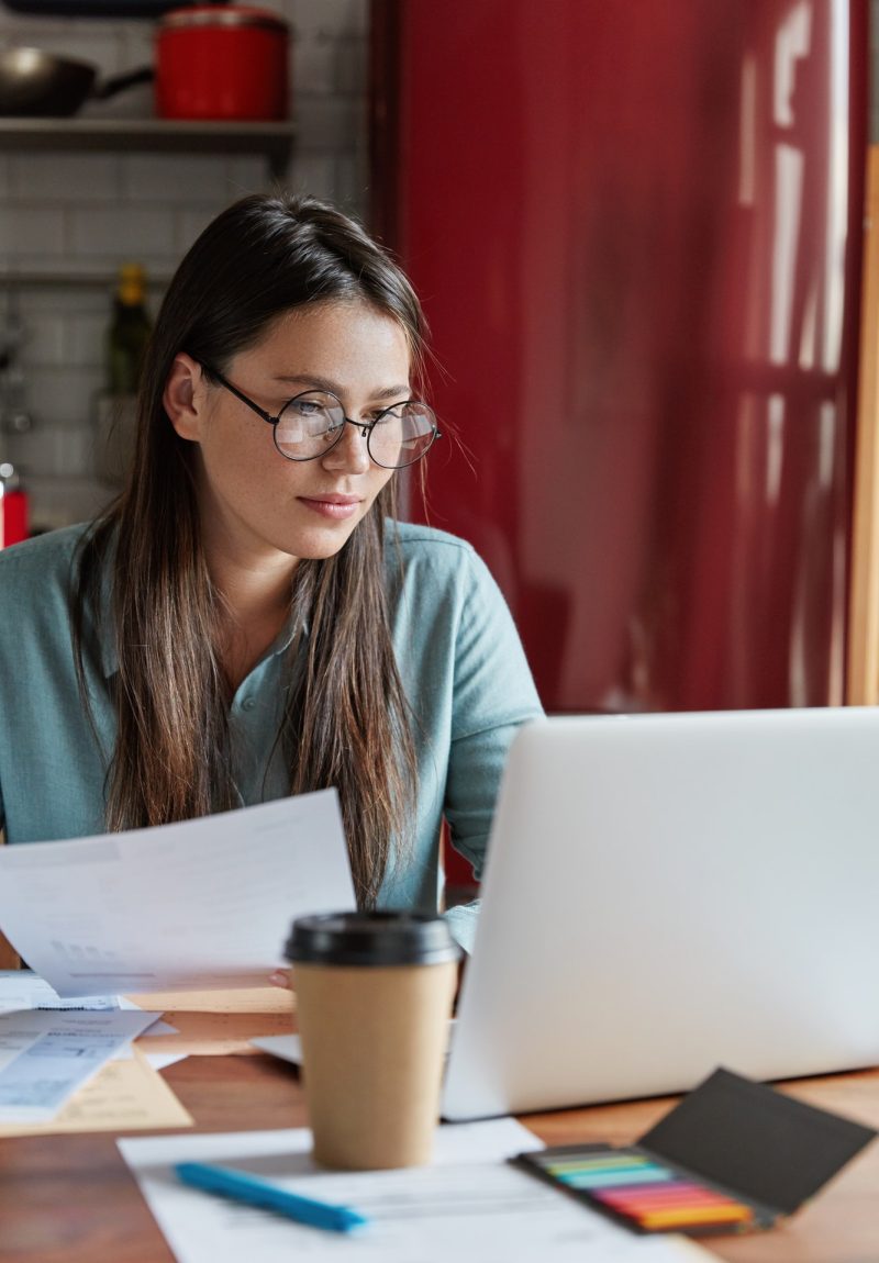Busy financier checks accountings from papers, focused into screen of laptop computer, wears round o