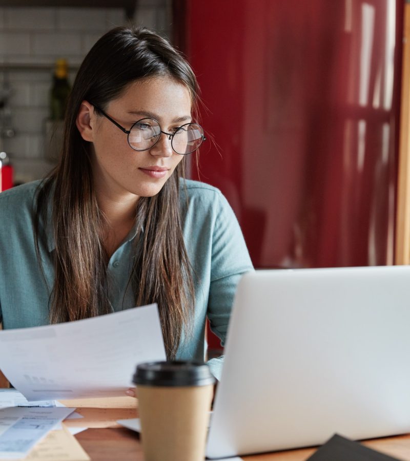 Busy financier checks accountings from papers, focused into screen of laptop computer, wears round o