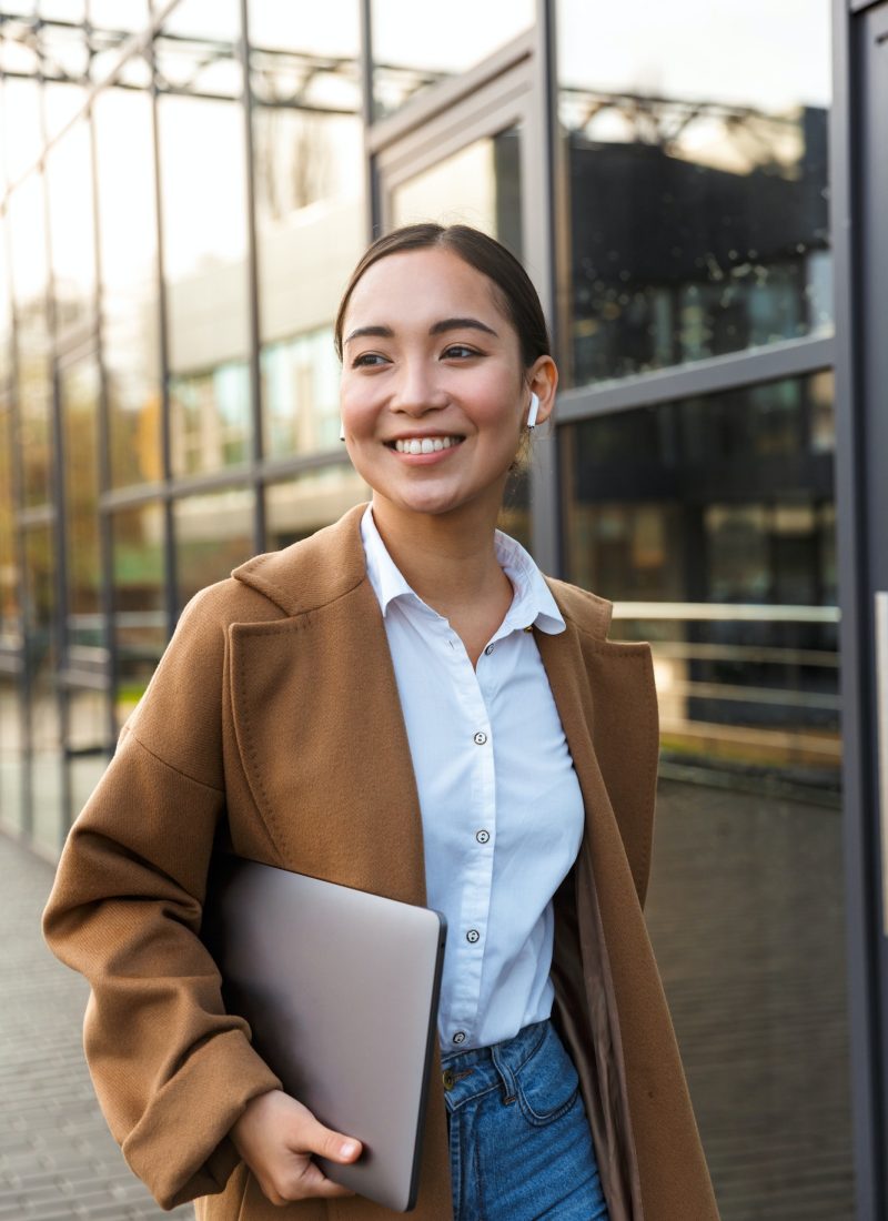 Office girl wearing a brown coat and white shirt, holding a laptop while walking on the street