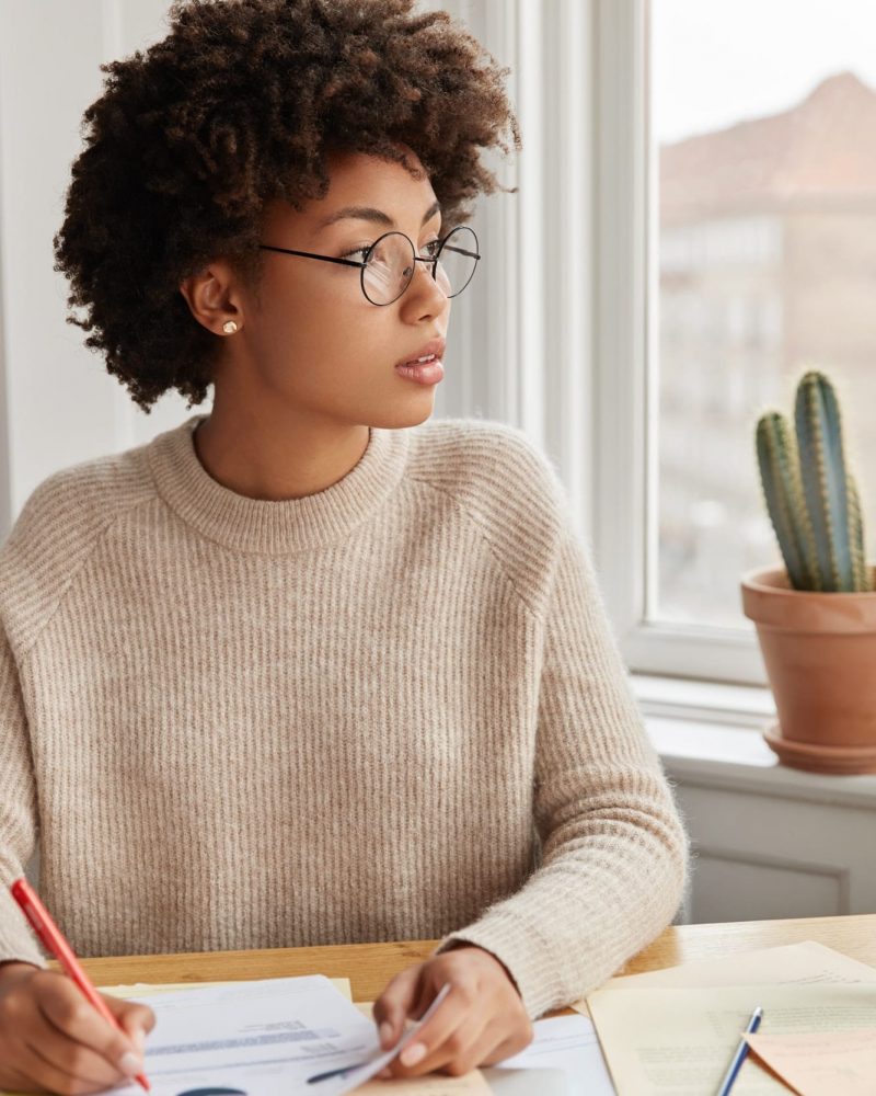 Pensive black businesswoman makes notes in documents, sits at desktop at modern apartment, dressed i
