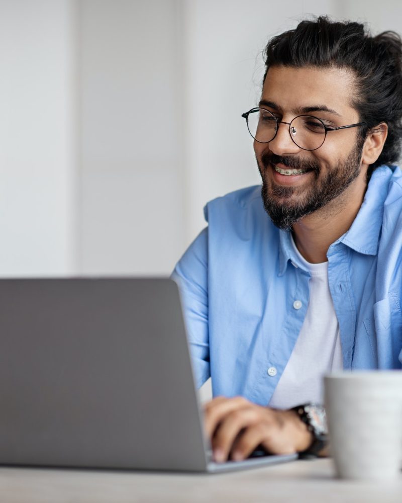 Smiling Indian Writer Using Laptop Computer, Sitting At Desk In Home Office