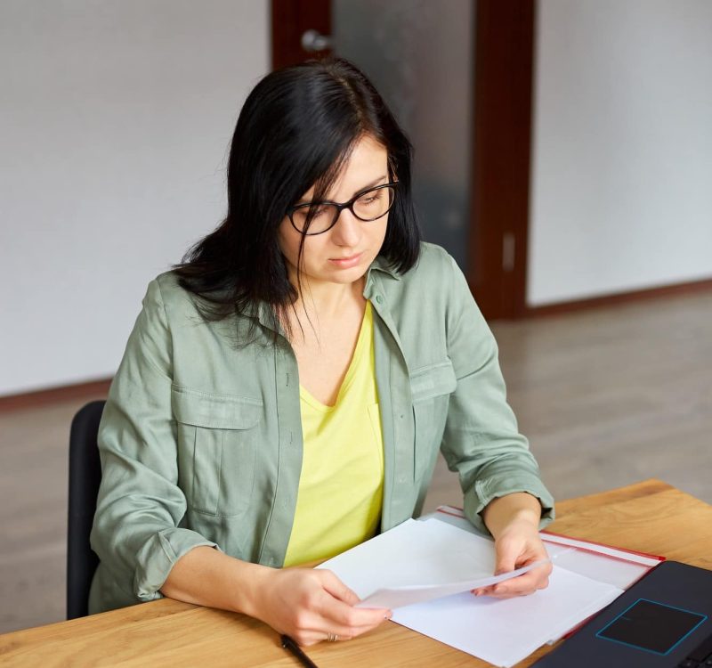 Stylish brunette woman in glasses sitting at wooden table with notepad working in her office