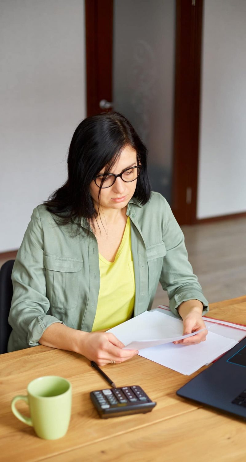 Stylish brunette woman in glasses sitting at wooden table with notepad working in her office