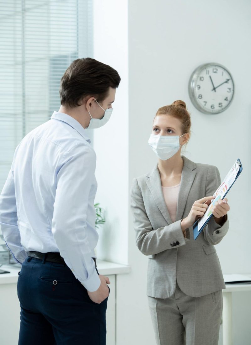 Two young elegant accountants in protective masks discussing financial document