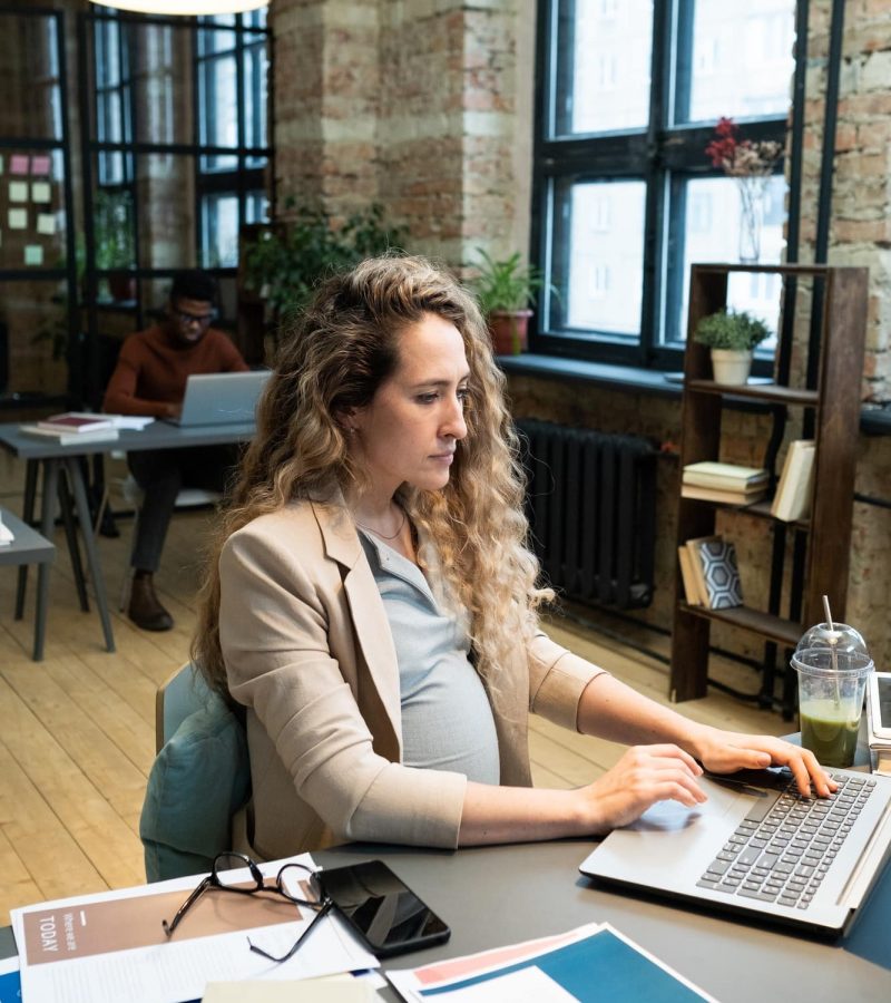 Young serious pregnant accountant sitting by desk in front of laptop